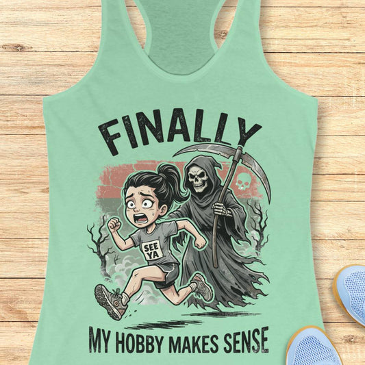 A mint green racerback tank top laid flat on a light wood background, featuring a cartoon graphic of a girl running toward a grim reaper with the text 'FINALLY MY HOBBY MAKES SENSE'.