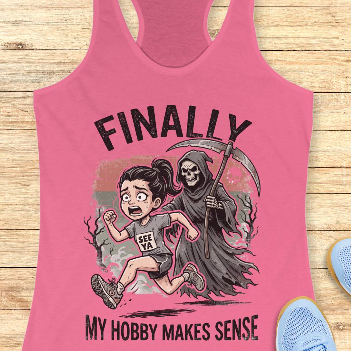 A pink racerback tank top laid flat on a light wood surface, featuring a cartoon graphic of a girl running and a grim reaper behind her with the text 'FINALLY MY HOBBY MAKES SENSE.' Partial blue shoes are visible at the edges as styling props.