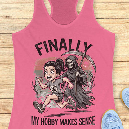 A pink racerback tank top laid flat on a light wood surface, featuring a cartoon graphic of a girl running and a grim reaper behind her with the text 'FINALLY MY HOBBY MAKES SENSE.' Partial blue shoes are visible at the edges as styling props.