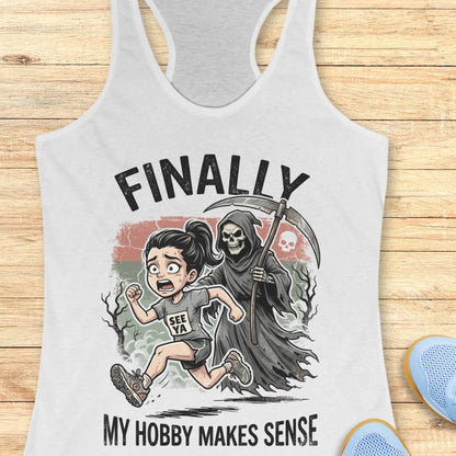 A white racerback tank top laid flat on a light wood background, featuring a cartoon graphic of a girl running while being chased by a grim reaper, with the text 'FINALLY MY HOBBY MAKES SENSE'.