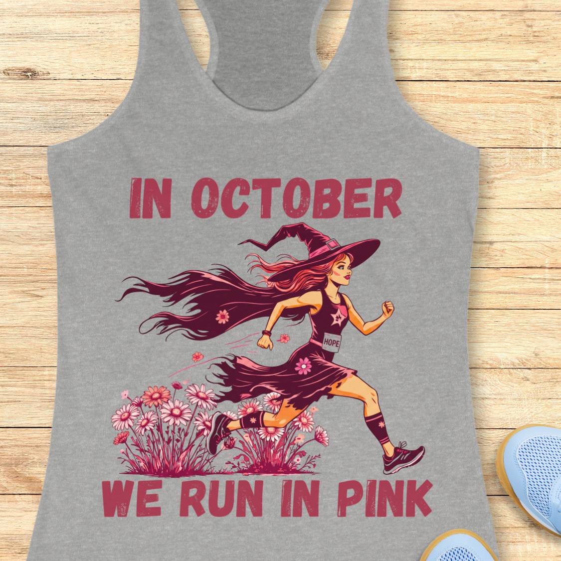 A heather gray racerback tank top laid flat on a light wood surface, featuring a graphic of a running witch with pink flowers and the text 'IN OCTOBER WE RUN IN PINK' in pink lettering.