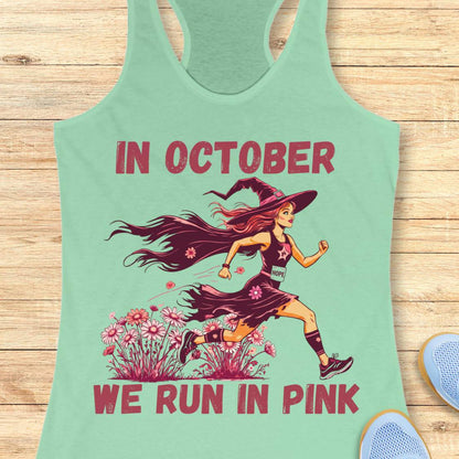 A mint green racerback tank top laid flat on a light wood background, featuring a graphic of a woman running in a witch hat with pink flowers and the text 'IN OCTOBER WE RUN IN PINK'.