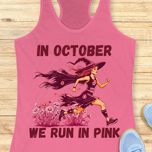 A pink racerback tank top laid flat on a light wood surface, featuring a graphic of a witch running through flowers with the text 'IN OCTOBER WE RUN IN PINK.' Partial blue shoes are visible at the lower right as styling props.