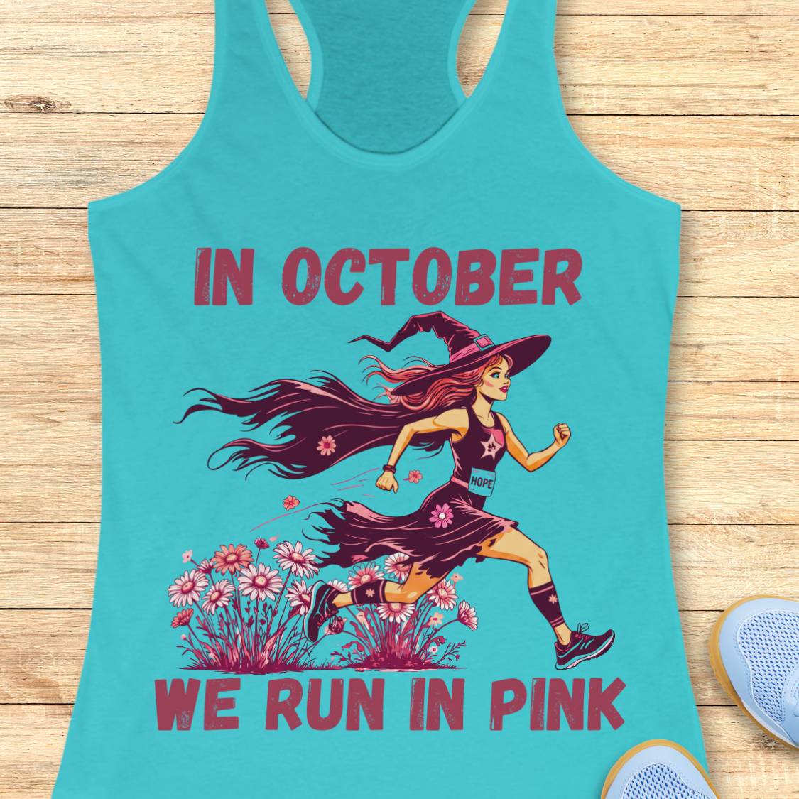 A turquoise racerback tank top laid flat on a light wood surface, featuring a graphic of a woman running in pink and purple tones with the text 'IN OCTOBER WE RUN IN PINK.' Part of a light blue shoe is visible at the lower right as a styling prop.