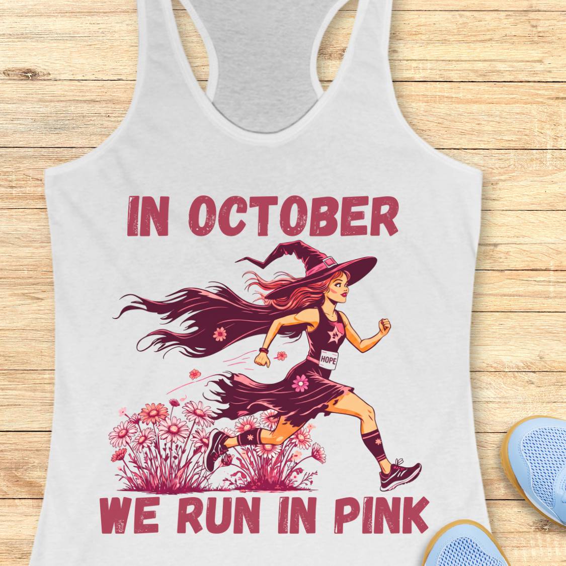 A white racerback tank top laid flat on a light wood surface, featuring a graphic of a woman running as a witch with the text 'IN OCTOBER WE RUN IN PINK' in pink. Part of a blue shoe is visible at the bottom right.