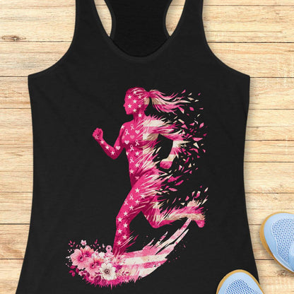 A black racerback tank top featuring a vibrant pink graphic of a running woman made of stars, pink ribbons and floral elements, laid flat on a light wood background with blue sneakers partially visible.