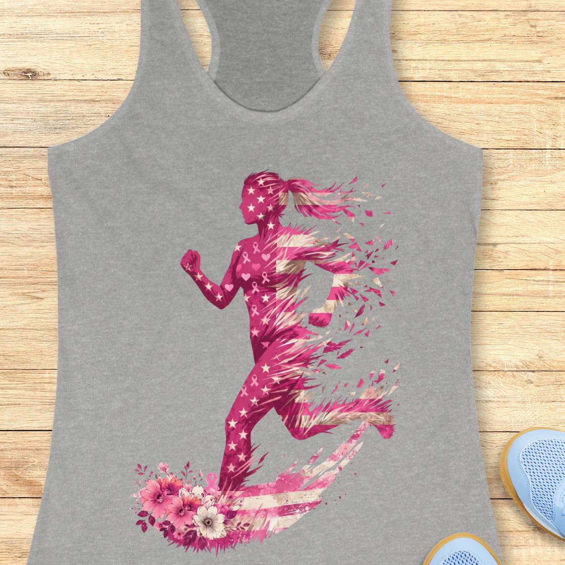 A heather gray racerback tank top featuring a vibrant pink and white graphic of a running woman with stars, pink ribbon and floral elements, displayed flat on a light wood background.