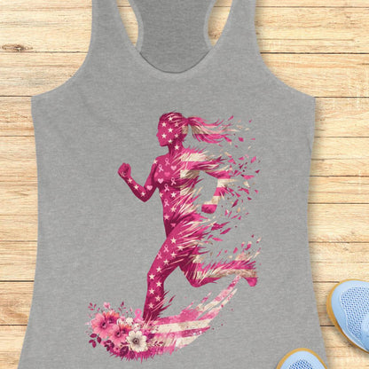 A heather gray racerback tank top featuring a vibrant pink and white graphic of a running woman with stars, pink ribbon and floral elements, displayed flat on a light wood background.