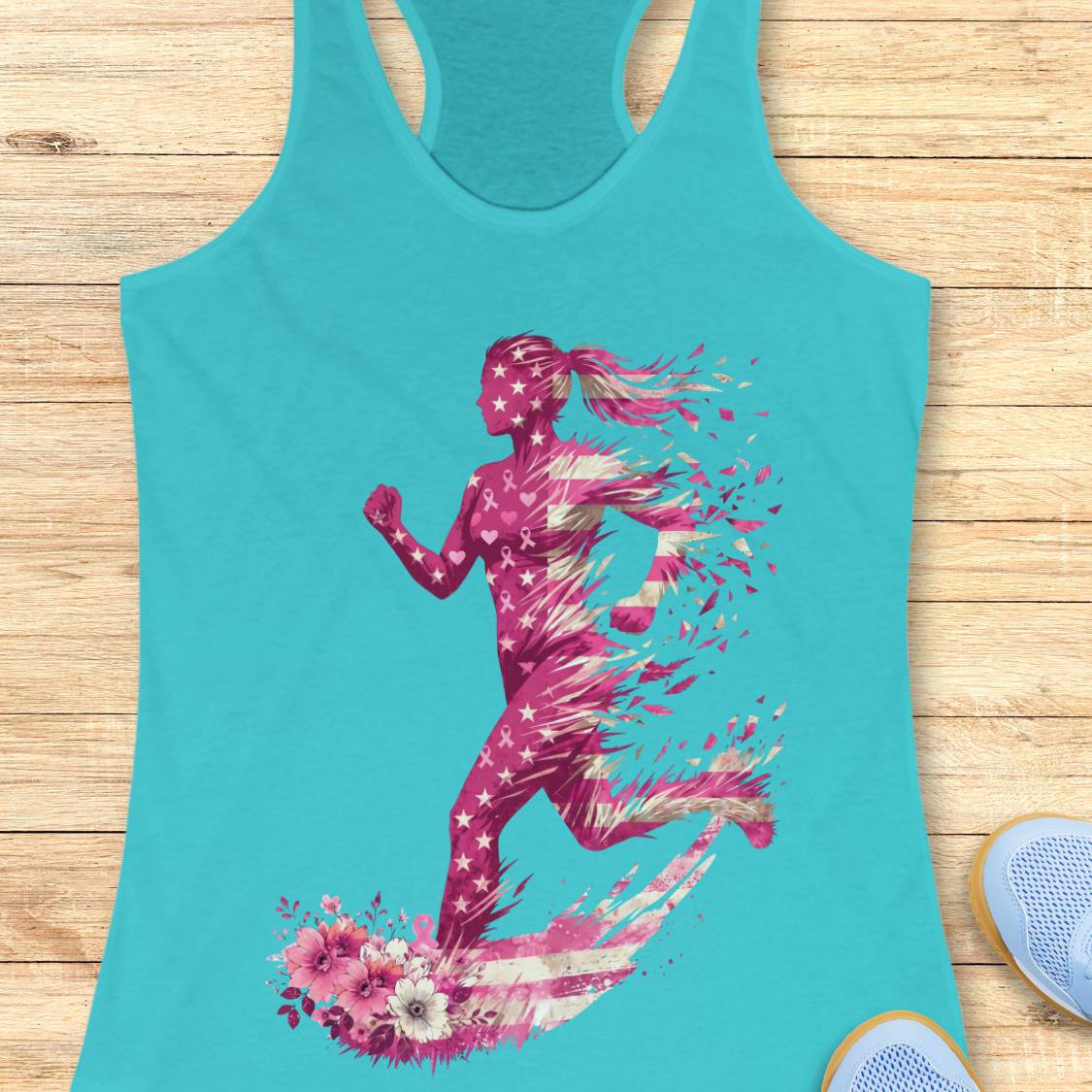 A turquoise racerback tank top laid flat on a light wood surface, featuring a large graphic of a running woman in pink and white with floral elements, and pink ribbons at the bottom. Partial blue sneakers are visible at the bottom right as styling props.