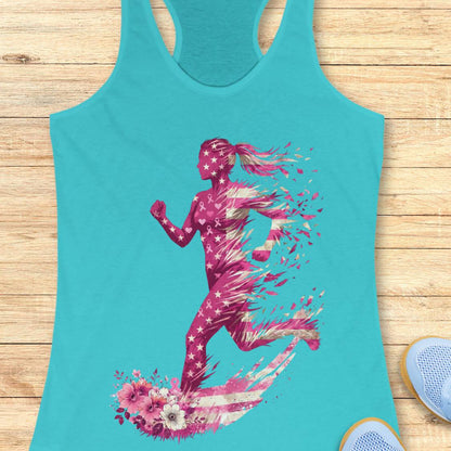 A turquoise racerback tank top laid flat on a light wood surface, featuring a large graphic of a running woman in pink and white with floral elements, and pink ribbons at the bottom. Partial blue sneakers are visible at the bottom right as styling props.