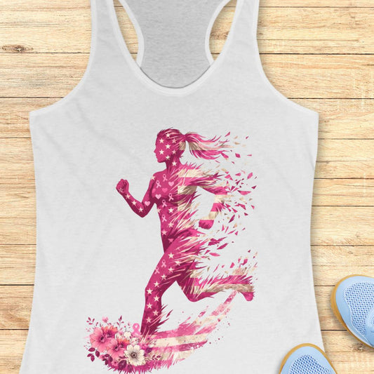 A white racerback tank top featuring a vibrant pink ribbon watercolor graphic of a running woman with stars, laid flat on a light wood background with blue sneakers partially visible.