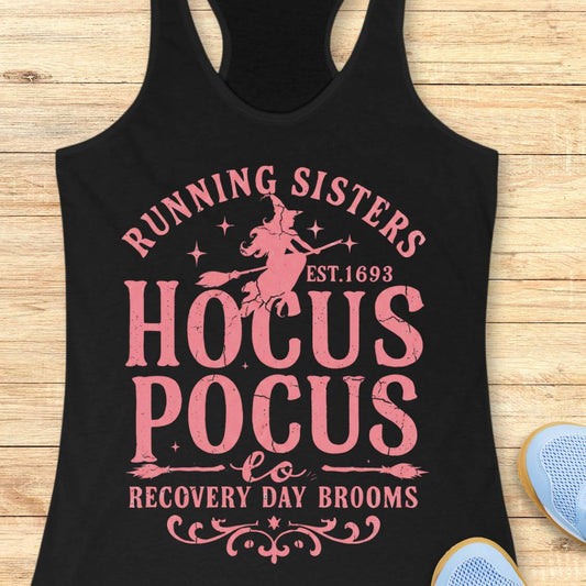 A black racerback tank top with a pink graphic print reading 'RUNNING SISTERS HOCUS POCUS RECOVERY DAY BROOMS' and a witch riding a broomstick, shown on a light wood background with partial blue shoes at the corner.