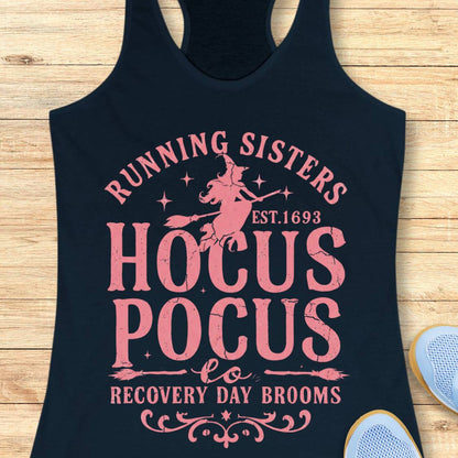 A navy racerback tank top with a pink distressed graphic reading 'Running Sisters Hocus Pocus to Recovery Day Brooms' with a witch riding a broomstick, laid flat on a light wood surface. Part of a blue shoe is visible at the bottom right.