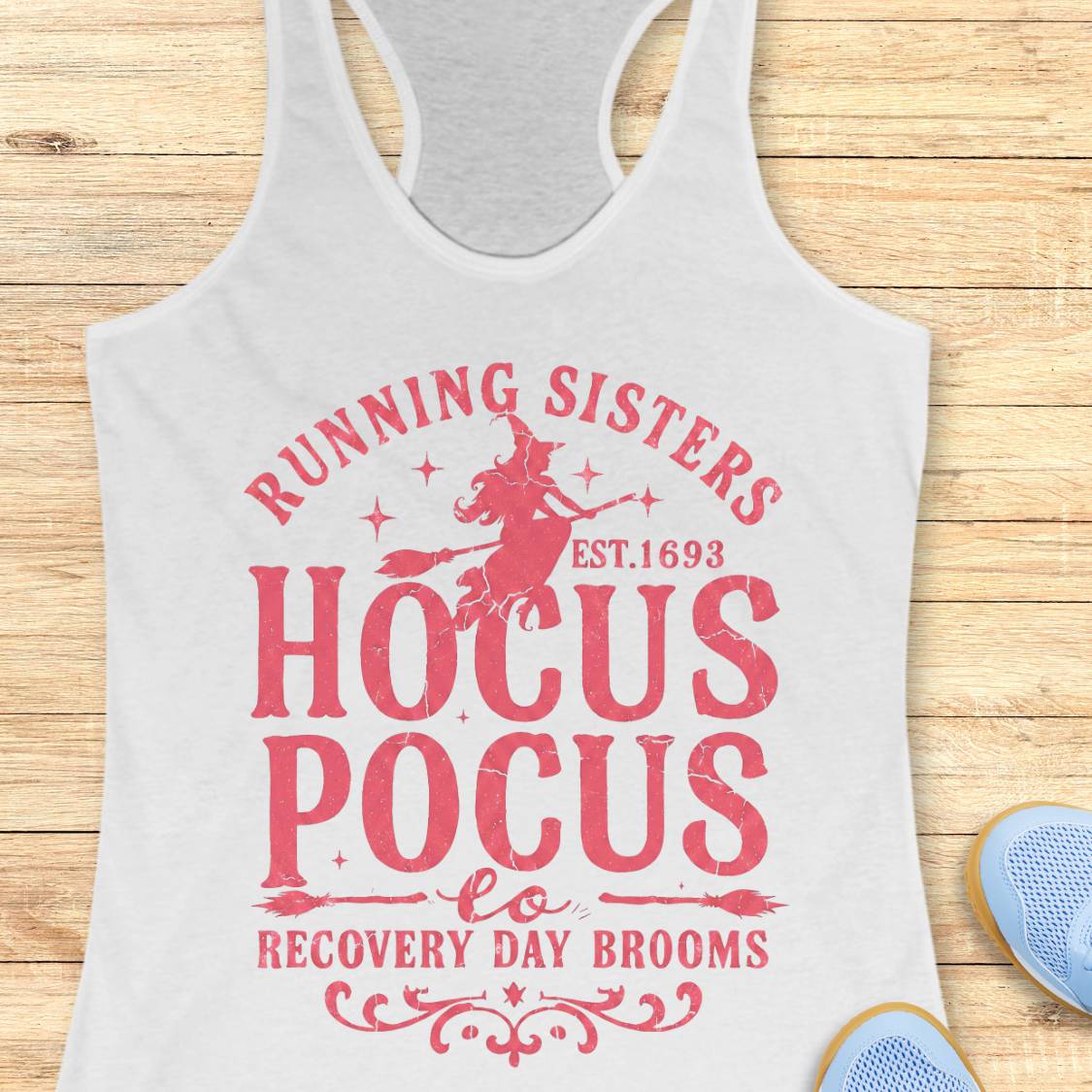 A white racerback tank top with a pink graphic print reading 'Running Sisters Hocus Pocus Recovery Day Brooms' with a witch riding a broom, shown on a wooden background with partial blue shoes at the bottom right.