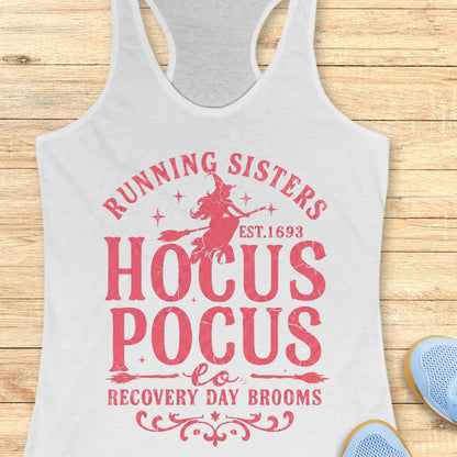 A white racerback tank top with a pink graphic print reading 'Running Sisters Hocus Pocus Recovery Day Brooms' with a witch riding a broom, shown on a wooden background with partial blue shoes at the bottom right.
