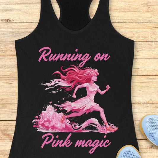 A black racerback tank top laid flat on a light wood surface, featuring a pink graphic of a running woman with the words 'Running on Pink magic' in pink script. Part of a blue shoe is visible at the bottom right as a styling prop.