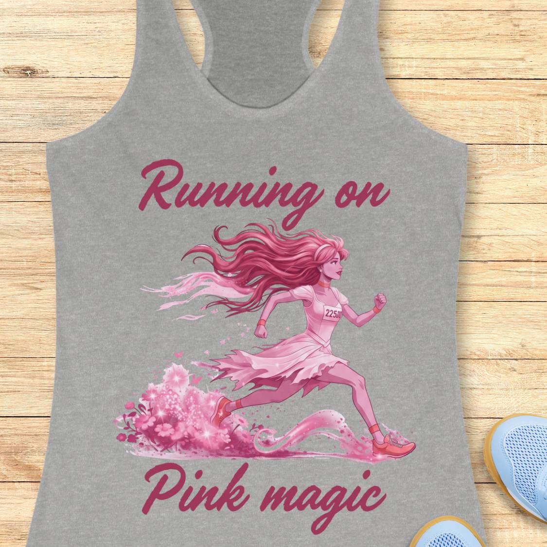 A heather gray racerback tank top laid flat on a light wood background, featuring a pink graphic of a running girl with the text 'Running on Pink magic.' Partial blue shoes are visible at the bottom right as styling props.