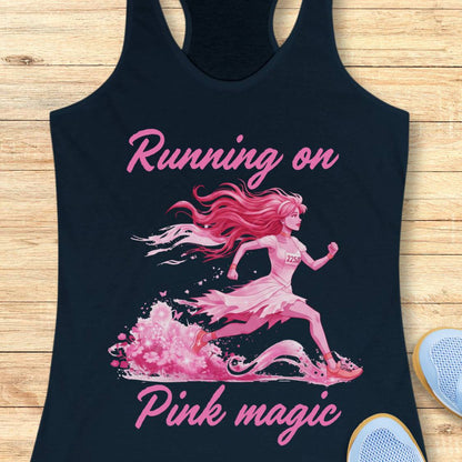 A navy racerback tank top laid flat on a light wood surface, featuring a pink graphic of a running girl with the words 'Running on Pink magic.' Part of a white sneaker is visible at the lower right.