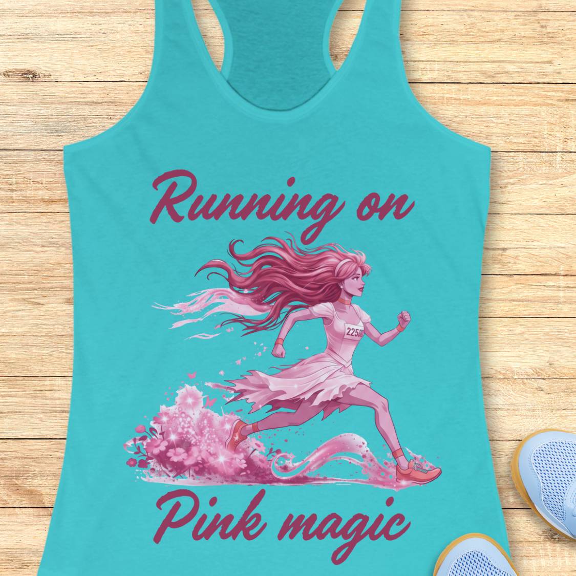 A turquoise racerback tank top laid flat on a light wood surface, featuring a pink graphic of a running woman with the text 'Running on Pink magic.' A light blue shoe is partially visible at the bottom right as a styling prop.