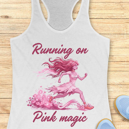 A white racerback tank top laid flat on a light wood background, featuring a pink graphic of a running girl and the text 'Running on Pink magic.' Partial blue shoes are visible at the bottom right as styling props.