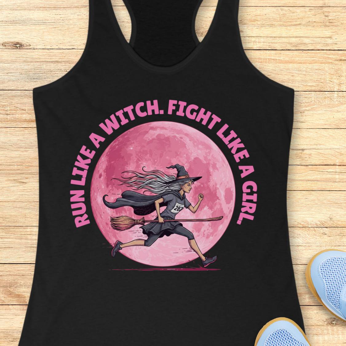 A black racerback tank top laid flat on a light wood background, featuring a pink graphic of a witch running on a broomstick in front of a large pink moon, with the text 'RUN LIKE A WITCH, FIGHT LIKE A GIRL' in pink around the design.