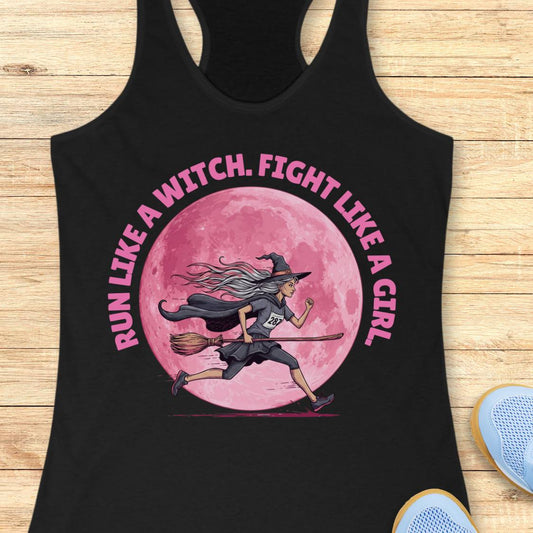 A black racerback tank top laid flat on a light wood background, featuring a pink graphic of a witch running on a broomstick in front of a large pink moon, with the text 'RUN LIKE A WITCH, FIGHT LIKE A GIRL' in pink around the design.