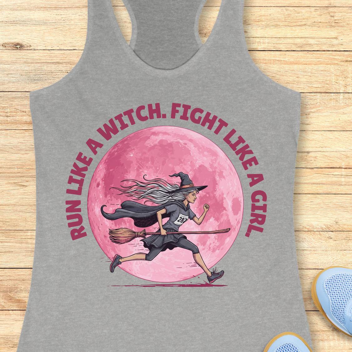 A heather gray racerback tank top laid flat on a light wood surface, featuring a large graphic print of a witch running on a broomstick in front of a pink full moon, with the text 'RUN LIKE A WITCH. FIGHT LIKE A GIRL' in pink around the design.