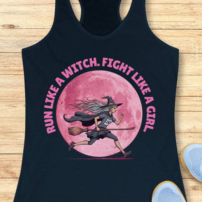 A navy racerback tank top laid flat on a light wood surface, featuring a large graphic print of a witch running on a broomstick in front of a pink full moon, with the text 'RUN LIKE A WITCH, FIGHT LIKE A GIRL' in pink around the design. Part of a white shoe is visible at the bottom right.