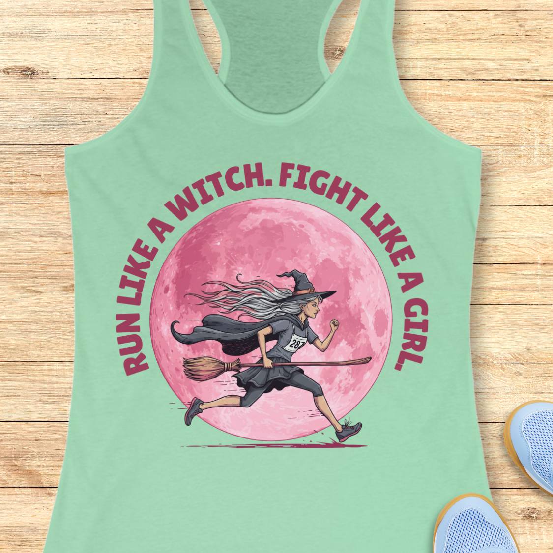 A mint green racerback tank top laid flat on a light wood background, featuring a large graphic of a witch running on a broomstick in front of a pink moon, with the text 'RUN LIKE A WITCH, FIGHT LIKE A GIRL' in pink around the design.