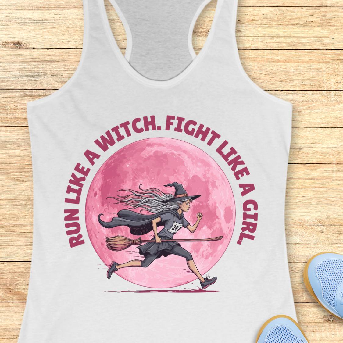 A white racerback tank top laid flat on a light wood surface, featuring a large graphic of a witch riding a broomstick in front of a pink moon, with the text 'RUN LIKE A WITCH, FIGHT LIKE A GIRL' in pink around the illustration.