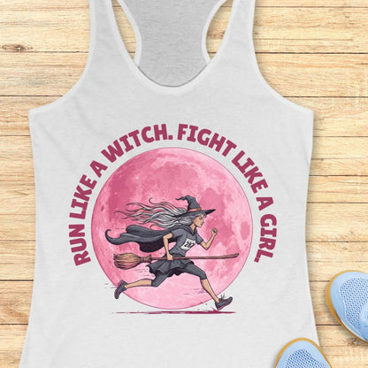 A white racerback tank top laid flat on a light wood surface, featuring a large graphic of a witch riding a broomstick in front of a pink moon, with the text 'RUN LIKE A WITCH, FIGHT LIKE A GIRL' in pink around the illustration.