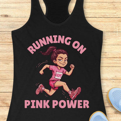 A black racerback tank top laid flat on a light wood background, featuring a cartoon illustration of a smiling female runner in a pink outfit with the words 'RUNNING ON PINK POWER' in pink text.
