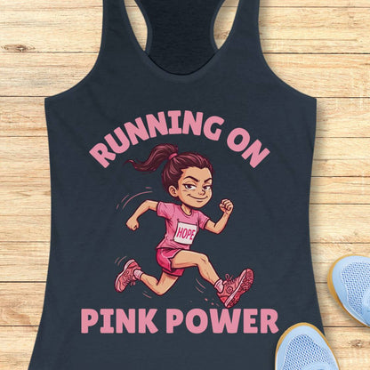 A indigo racerback tank top laid flat on a light wood surface, featuring a pink graphic of a running cartoon girl with the words 'RUNNING ON PINK POWER.' Part of a blue shoe is visible at the lower right as a styling prop.