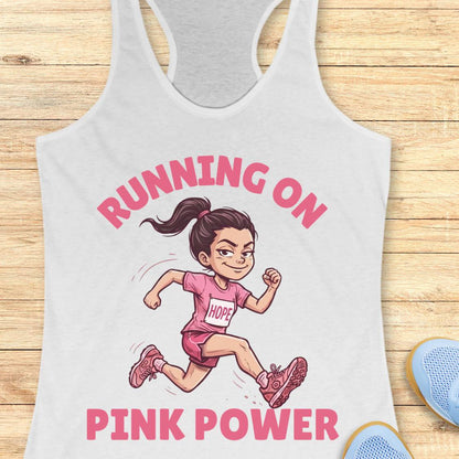 A white racerback tank top laid flat on a light wood background, featuring a cartoon illustration of a girl running with the text 'RUNNING ON PINK POWER' in pink. Part of a blue shoe is visible at the bottom right as a styling prop.