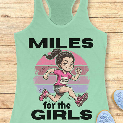 A mint green racerback tank top laid flat on a light wood background, featuring a cartoon illustration of a girl running with the text 'MILES for the GIRLS' in bold black letters.