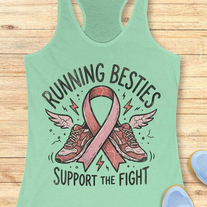A mint green racerback tank top laid flat on a light wood background, featuring a graphic print of a pink ribbon, running shoes, wings, and the text 'RUNNING BESTIES SUPPORT THE FIGHT'.