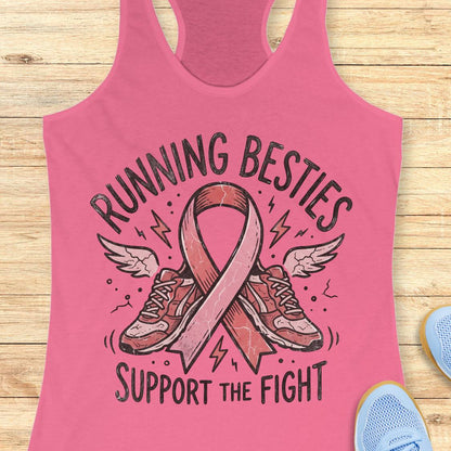 A pink racerback tank top laid flat on a light wood surface, featuring a graphic with the words 'RUNNING BESTIES SUPPORT THE FIGHT' around an illustrated pink ribbon and running shoes. Partial blue shoes appear at the bottom right as styling props.