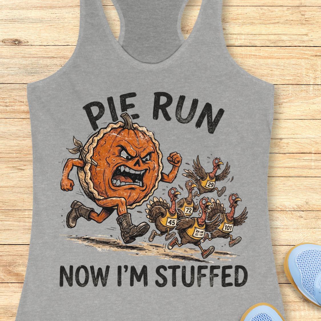 A heather gray racerback tank top laid flat on a light wood background, featuring a graphic print of an angry pie running and being chased by several cartoon turkeys. The text reads 'PIE RUN NOW I'M STUFFED'.