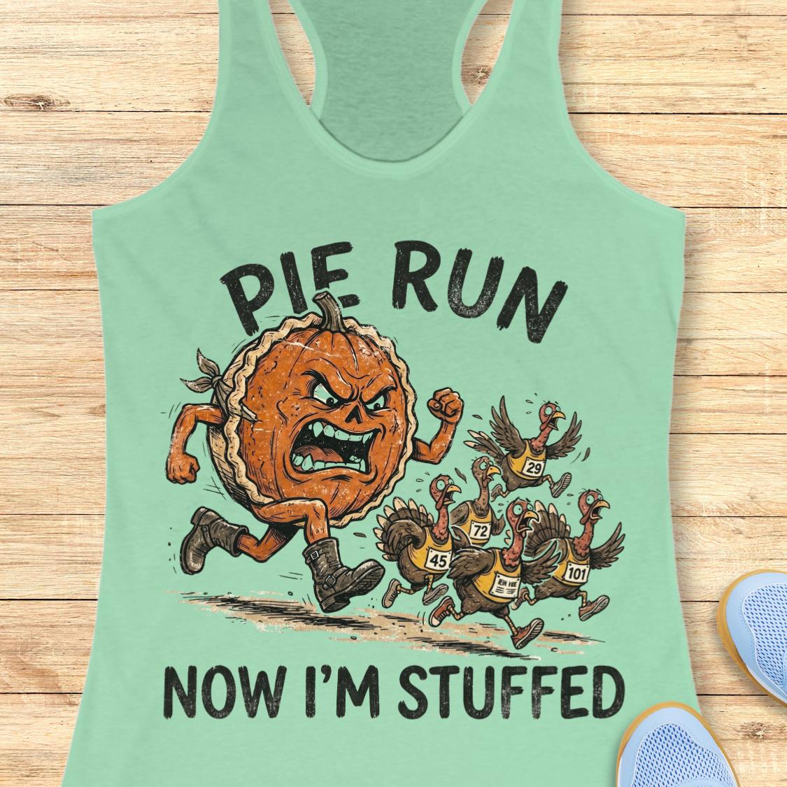 A mint green racerback tank top laid flat on a light wood background, featuring a graphic of an angry pie running after several turkeys with the text 'PIE RUN NOW I'M STUFFED'.