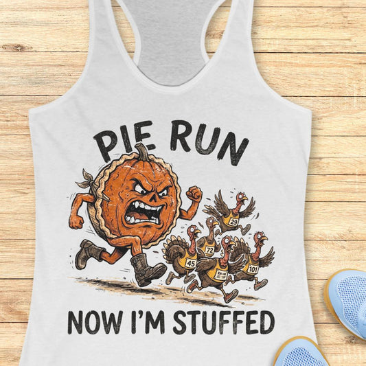 A white racerback tank top laid flat on a light wood background, featuring a graphic of an angry pie running like a race with several turkeys chasing it. The text reads 'PIE RUN NOW I'M STUFFED'.