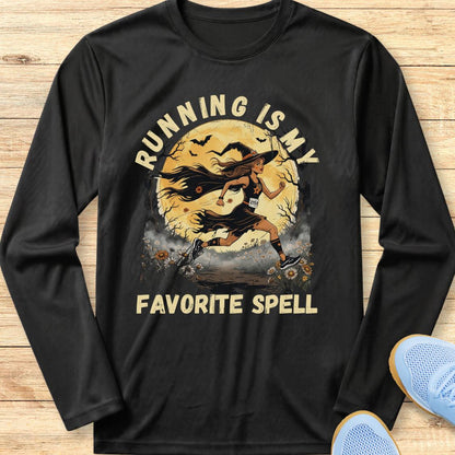 A black long-sleeve t-shirt with a graphic print of a witch running with a staff in front of a full moon, with the text 'RUNNING IS MY FAVORITE SPELL' in beige. The shirt is laid flat on a light wood background with blue sneakers partially visible in the corner.