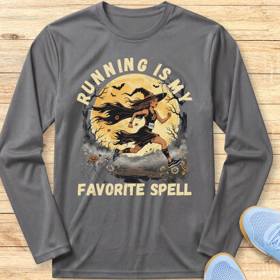 A dark gray long-sleeve t-shirt with a graphic of a female runner in front of a full moon, with the text 'RUNNING IS MY FAVORITE SPELL' printed on the front. The shirt is laid flat on a light wood background with a pair of blue sneakers partially visible in the corner.