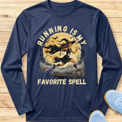 A navy blue long-sleeve t-shirt with a graphic print of a witch running in front of a full moon, with the text 'RUNNING IS MY FAVORITE SPELL' in beige. The shirt is laid flat on a light wood background with a pair of blue sneakers partially visible in the corner.