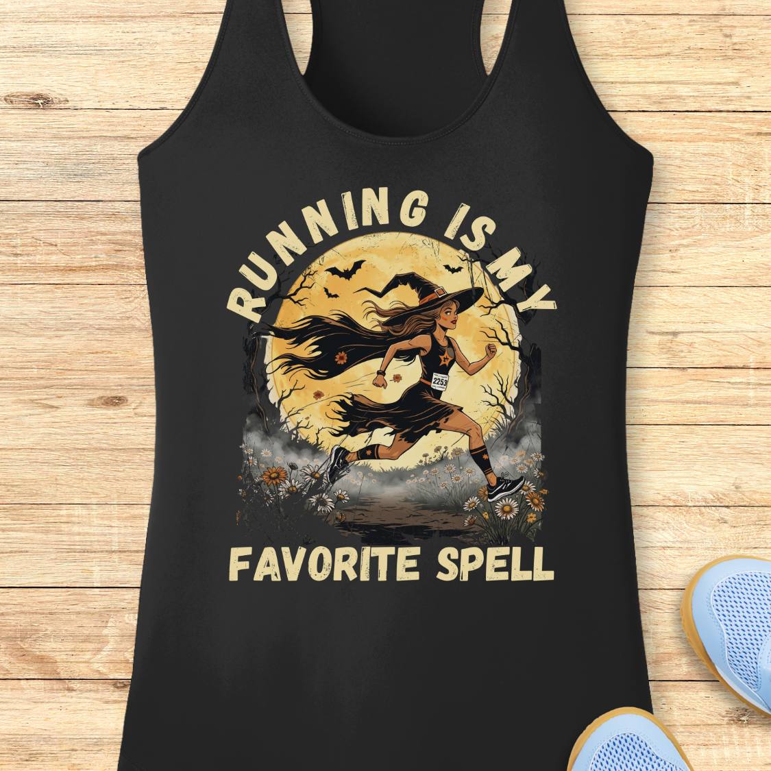 A black racerback tank top laid flat on a light wood background, featuring a distressed graphic of a female runner in front of a full moon with the text 'RUNNING IS MY FAVORITE SPELL'.