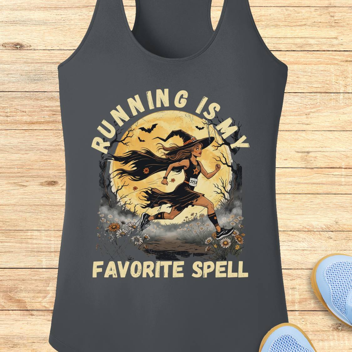 A dark gray racerback tank top with a graphic of a witch running in front of a full moon, with the text 'RUNNING IS MY FAVORITE SPELL' printed in beige. The tank top is displayed flat on a light wood background with a pair of blue sneakers partially visible in the corner.