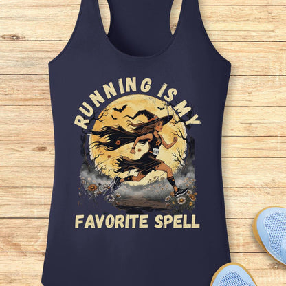 A navy blue racerback tank top with a graphic of a witch running in front of a full moon, with the text 'RUNNING IS MY FAVORITE SPELL' printed in beige. The tank top is displayed flat on a light wood background with a pair of blue sneakers partially visible in the corner.