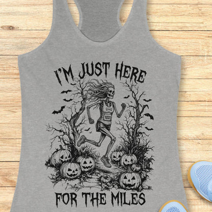 A heather gray racerback tank top laid flat on a light wood surface, featuring a black graphic of a skeleton runner surrounded by pumpkins and bats, with the text 'I'M JUST HERE FOR THE MILES'.