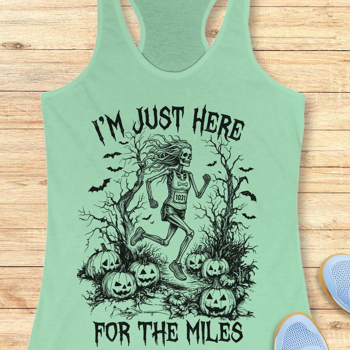 A mint green racerback tank top with a black graphic of a skeleton runner surrounded by pumpkins and bats, with the text 'I'M JUST HERE FOR THE MILES', displayed flat on a light wood background.