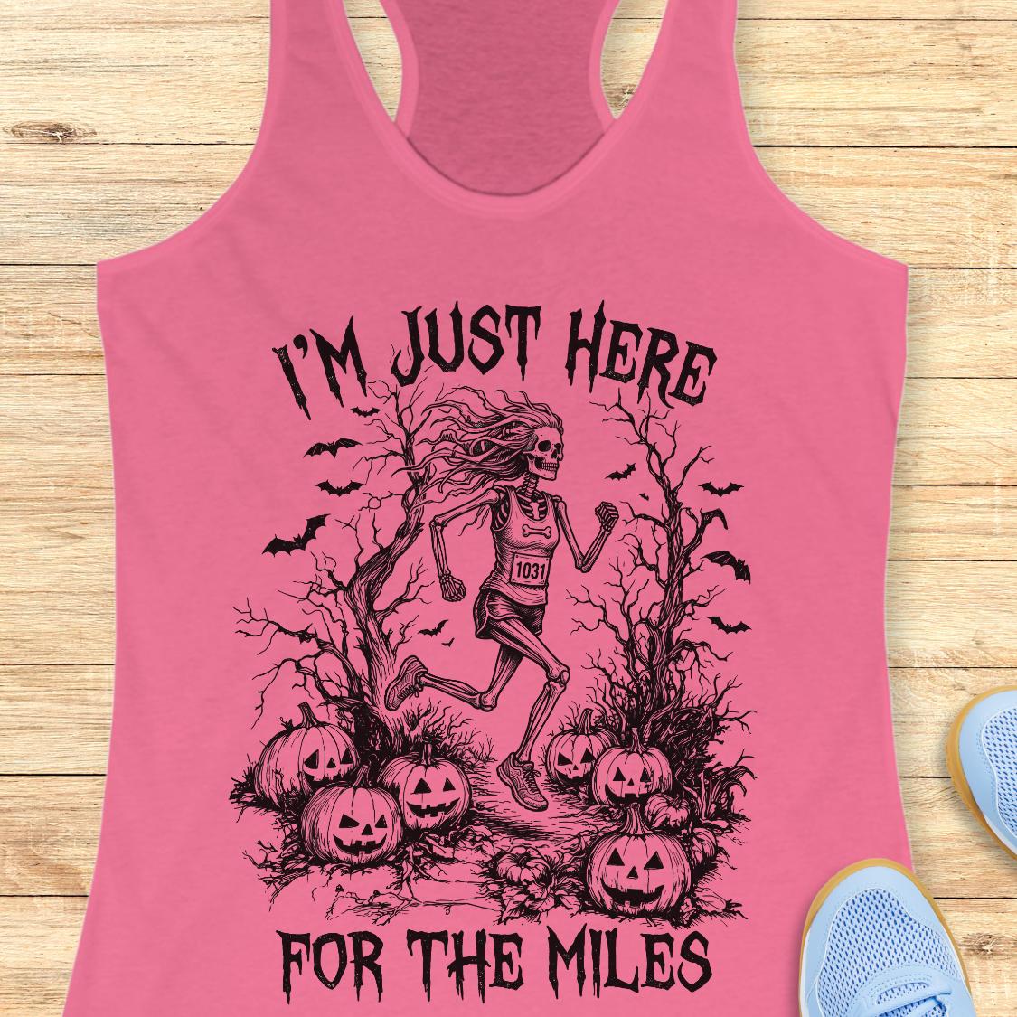 A pink racerback tank top laid flat on a light wood surface, featuring a black graphic of a skeleton runner surrounded by pumpkins, bats, and bare trees, with the text 'I'M JUST HERE FOR THE MILES'. Partial blue shoes are visible at the edges.