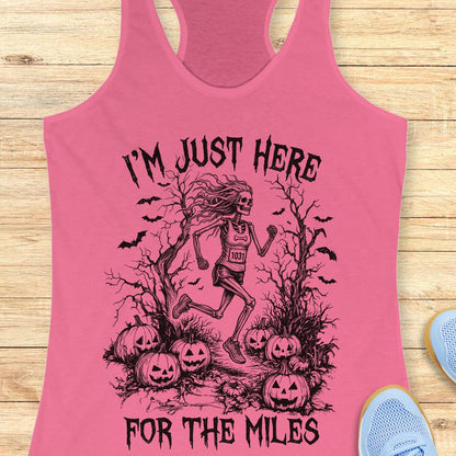 A pink racerback tank top laid flat on a light wood surface, featuring a black graphic of a skeleton runner surrounded by pumpkins, bats, and bare trees, with the text 'I'M JUST HERE FOR THE MILES'. Partial blue shoes are visible at the edges.